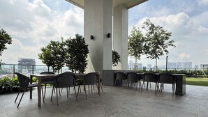 Un área de estar al aire libre con mesas y sillas, rodeada de grandes pilares y plantas en macetas, con vistas a un paisaje urbano.