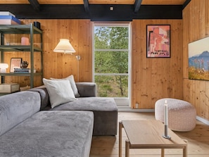 Ein gemütliches Wohnzimmer mit einer Couch, einem Tisch und einer Lampe. Ein Fenster bietet Blick auf Bäume, an der Wand hängt ein Poster.