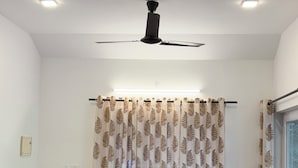 A room with a ceiling fan, a sofa with patterned cushions, two chairs, a small table, and a window with curtains.