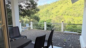 A covered outdoor seating area with a table and chairs, a view of a green hillside, and a railing.