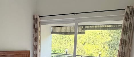 A bedroom with a bed, a ceiling fan, a window with a view of greenery, and a balcony with a railing.