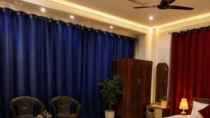 Habitación de hotel con cama, dos sillones, mesita de noche, lámpara, planta en maceta y ventilador de techo.