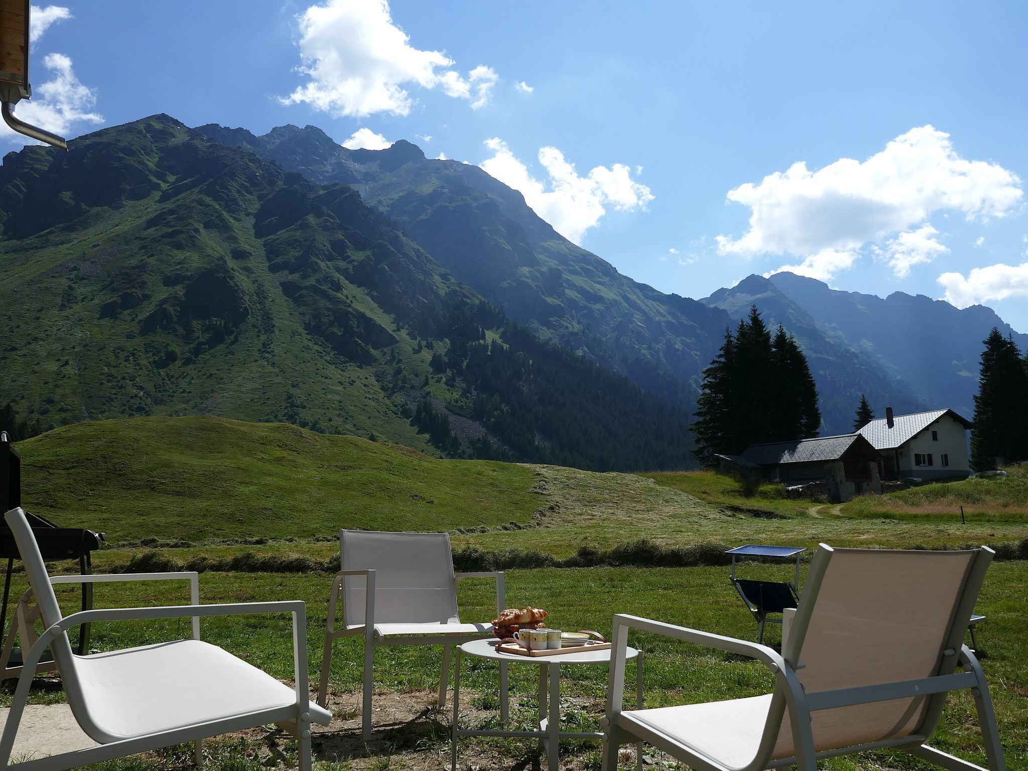 A table with chairs in a grassy area, overlooking a mountain range with a house in the background.