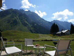 Ein Tisch mit Stühlen in einer grünen Wiese, mit Blick auf ein Gebirge, im Hintergrund ein Haus.