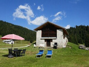 Ein Steinhaus mit geneigtem Dach, einer Terrasse mit Stühlen und Tisch sowie einem roten Sonnenschirm.