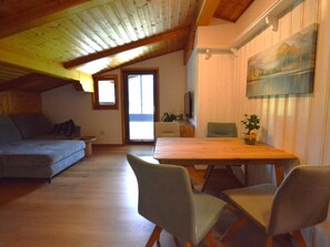 Ein gemütliches Loft-Zimmer mit Holzbalkendecke, einem Esstisch mit Stühlen, einer Couch und einem an der Wand befestigten Fernseher.