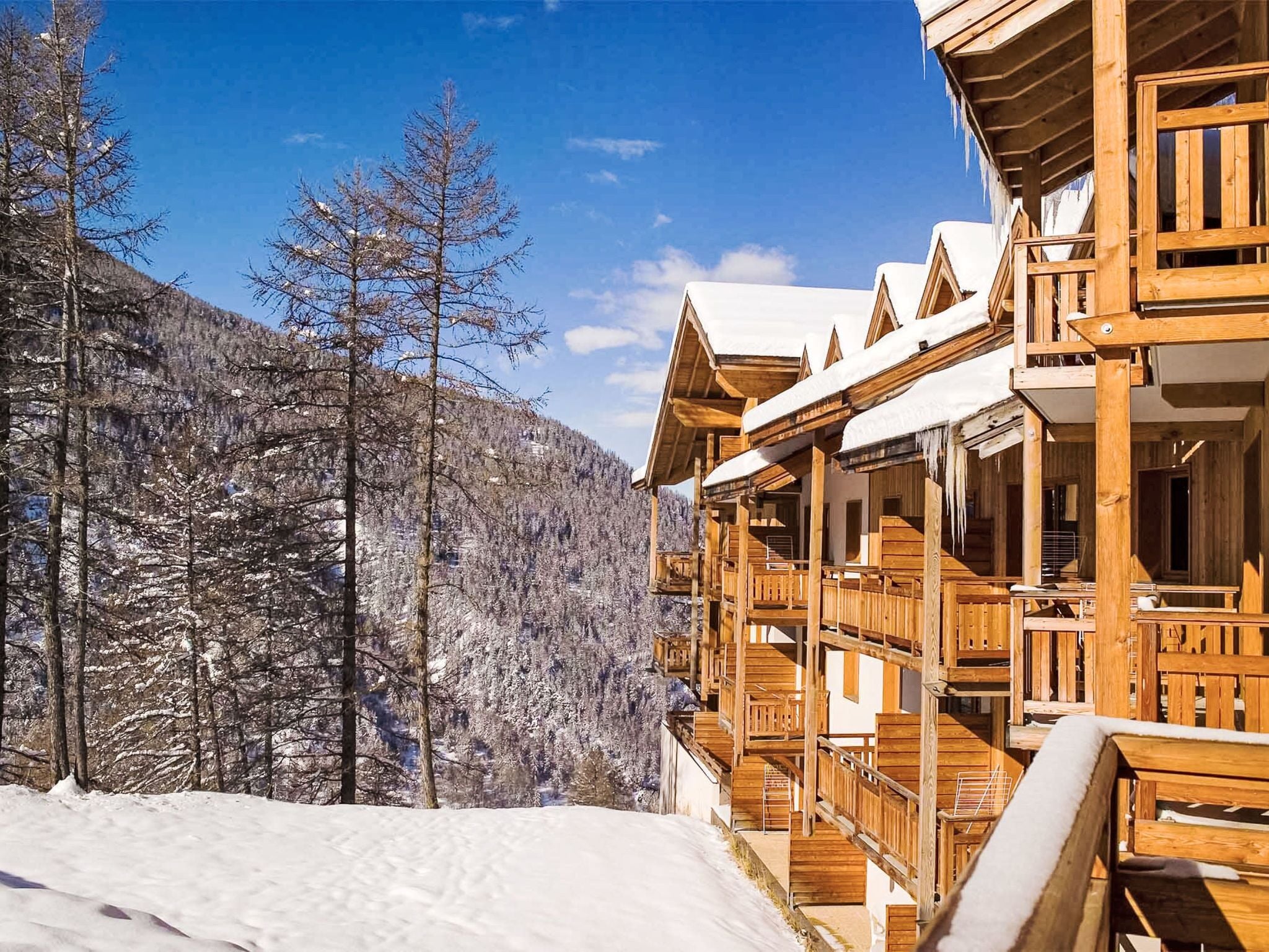 A wooden chalet with a balcony, surrounded by snow-covered trees and a mountain in the background.