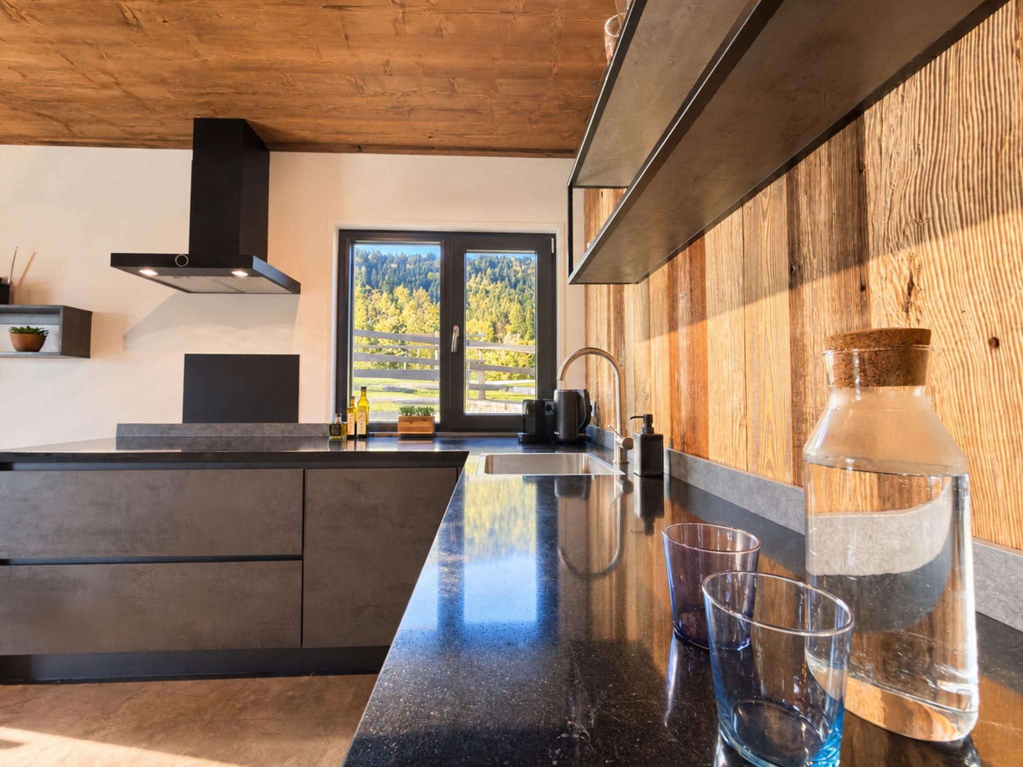 A modern kitchen with a large island, stainless steel appliances, and a view of a forest through a large window.