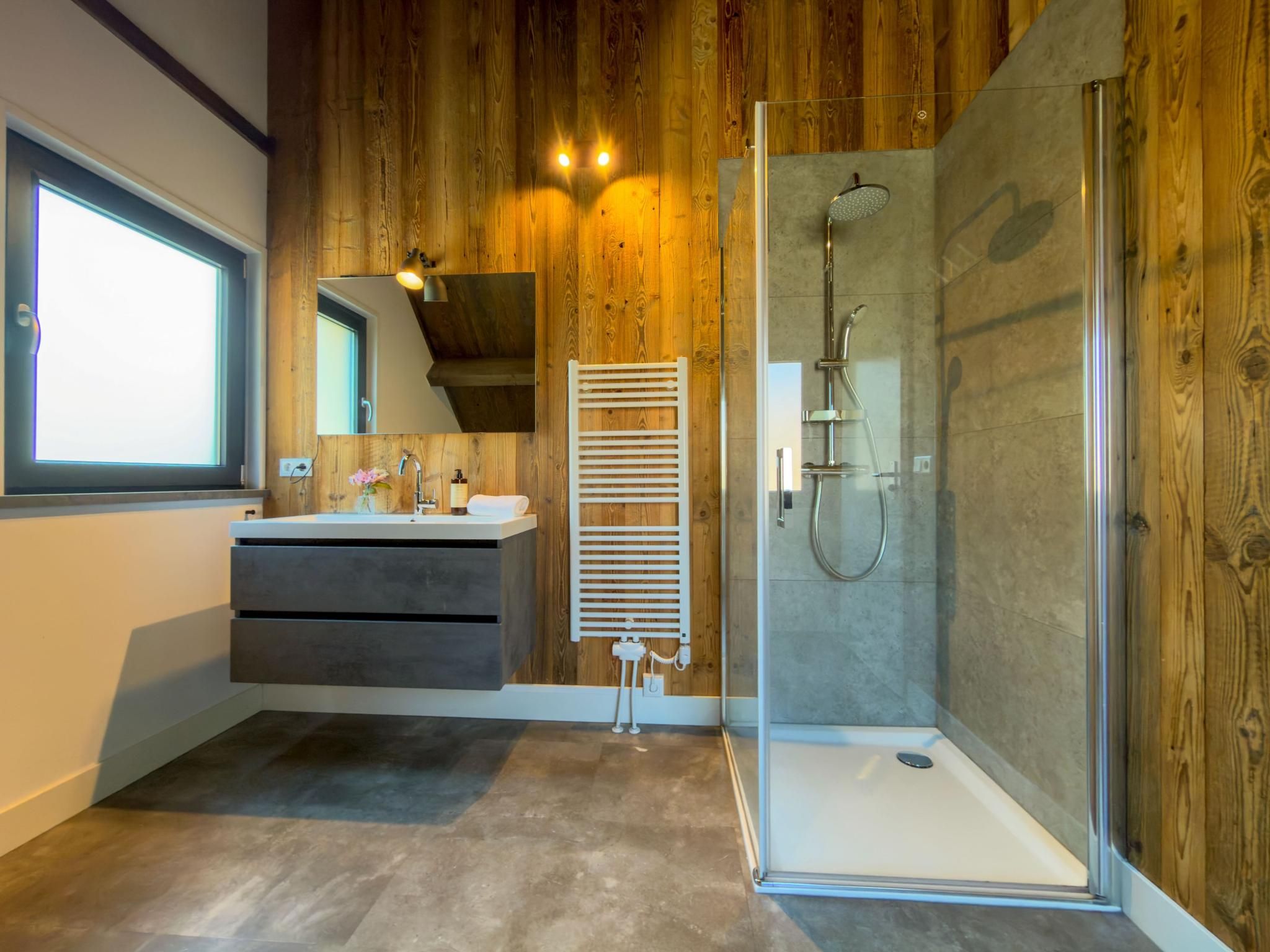 A modern bathroom with a glass-enclosed shower, wooden paneling, a vanity with a mirror, and a window.