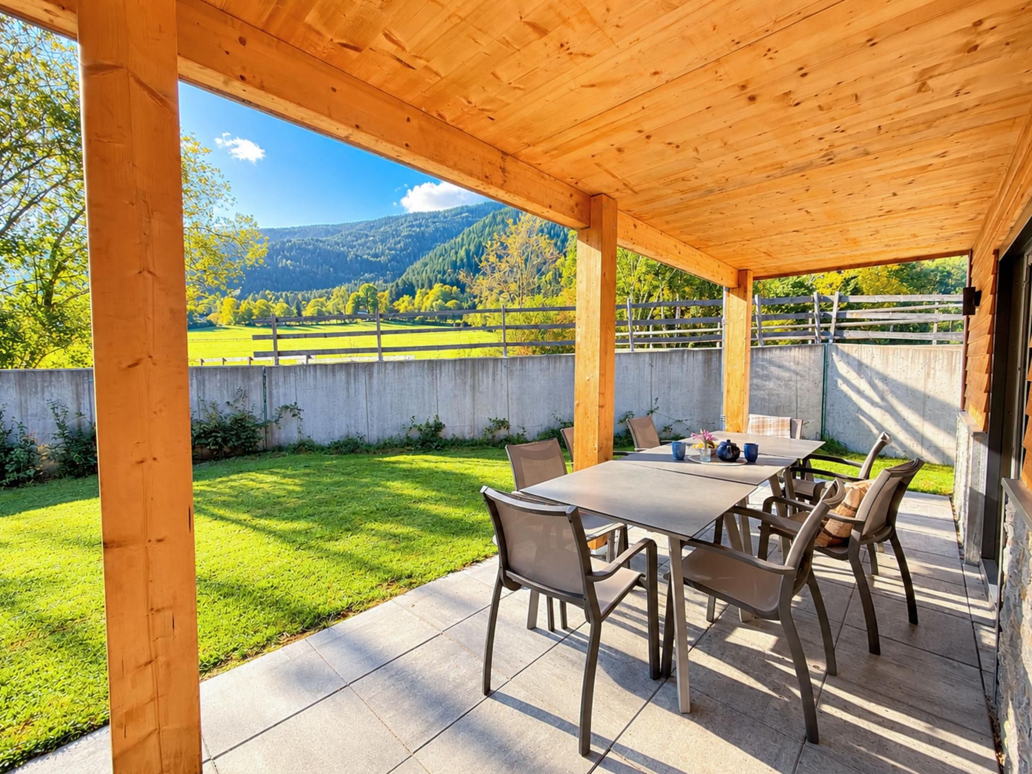 Une terrasse extérieure avec une table et des chaises, une vue sur un champ verdoyant et des montagnes, et une structure de pergola en bois.