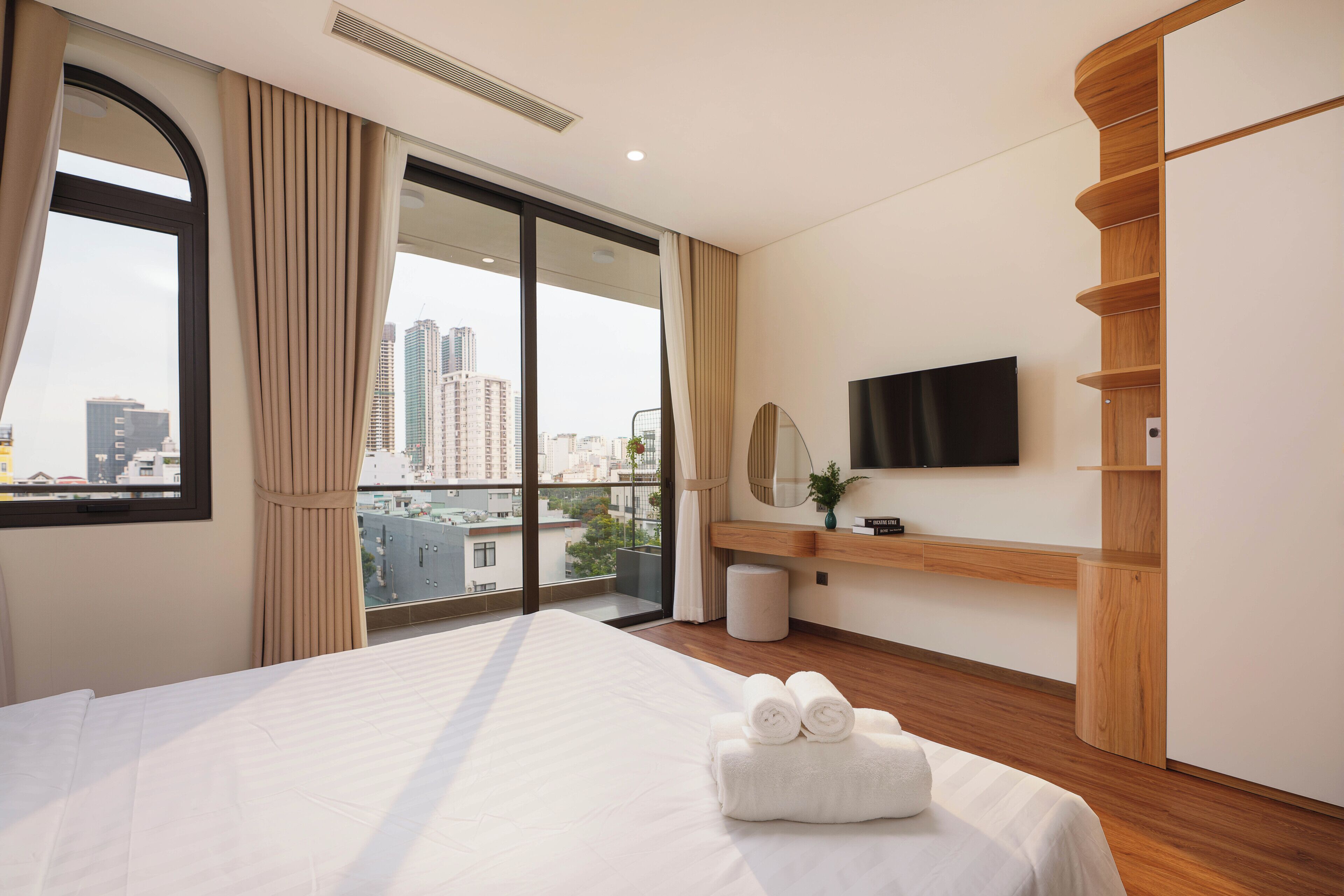 A hotel room with a large bed, a TV mounted on the wall, and a view of the cityscape through the window.