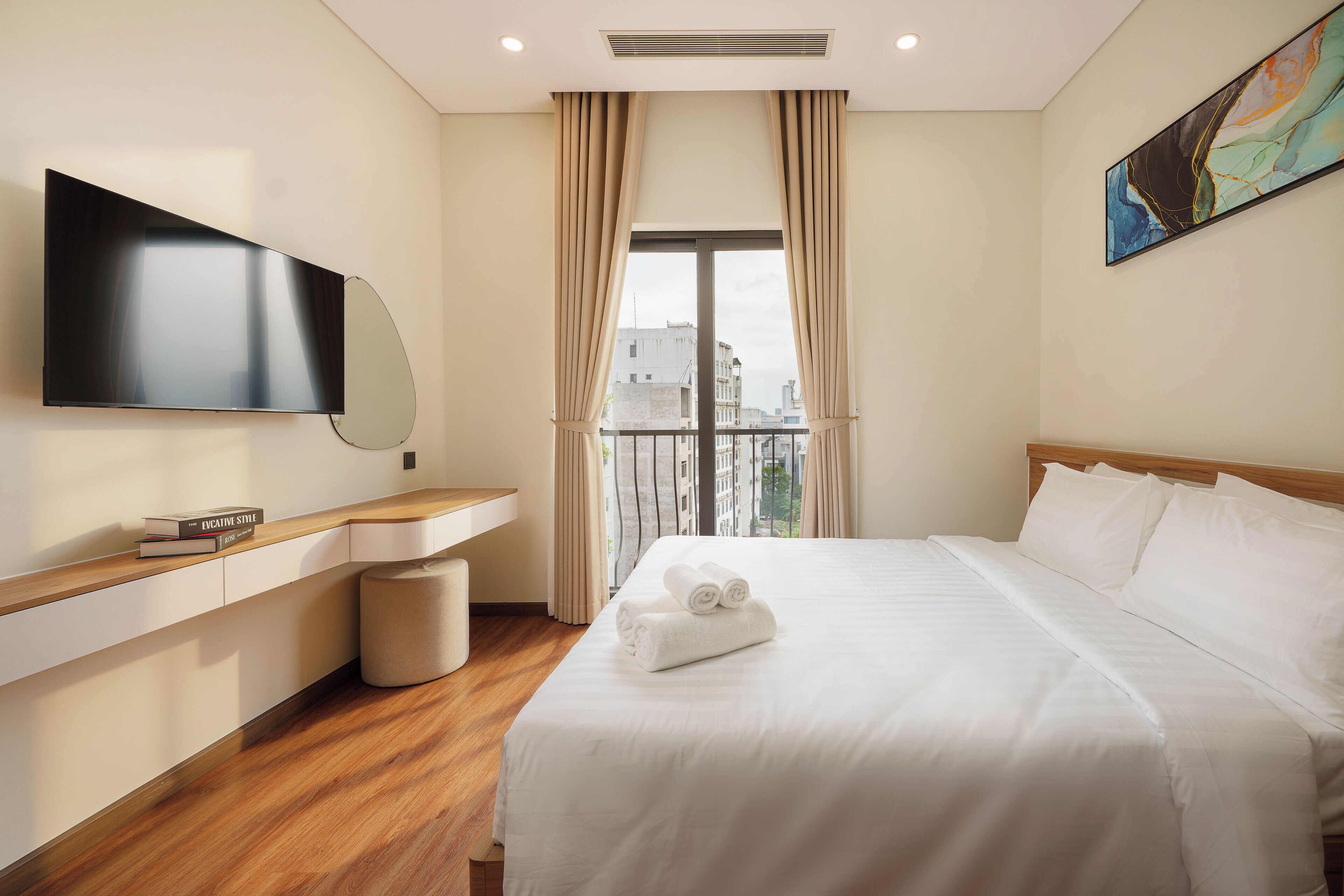 A hotel room with a bed, a TV, a bedside table, a window with curtains, and a view of buildings.