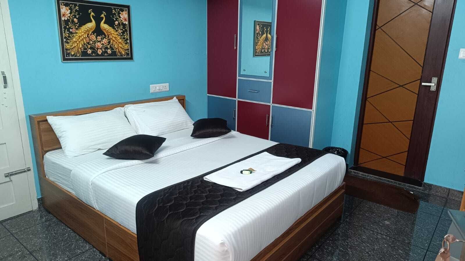 A neatly made bed with white linens and a black throw, a wooden headboard, and a blue wall with a decorative peacock painting.