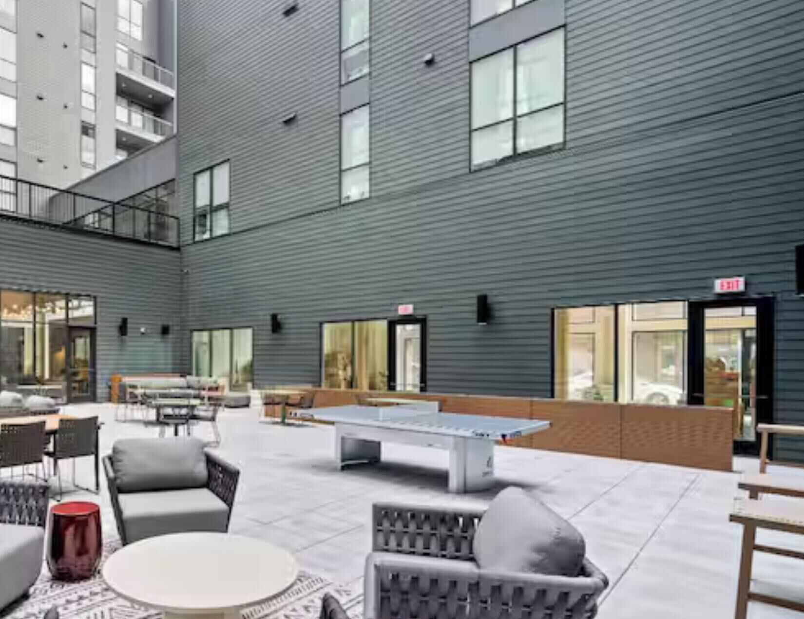 A modern outdoor seating area with tables and chairs, a ping pong table, and a building with large windows and an exit sign.