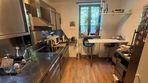 Cocina compacta con microondas, estufa y lavamanos. Se ve vegetación a través de la ventana.