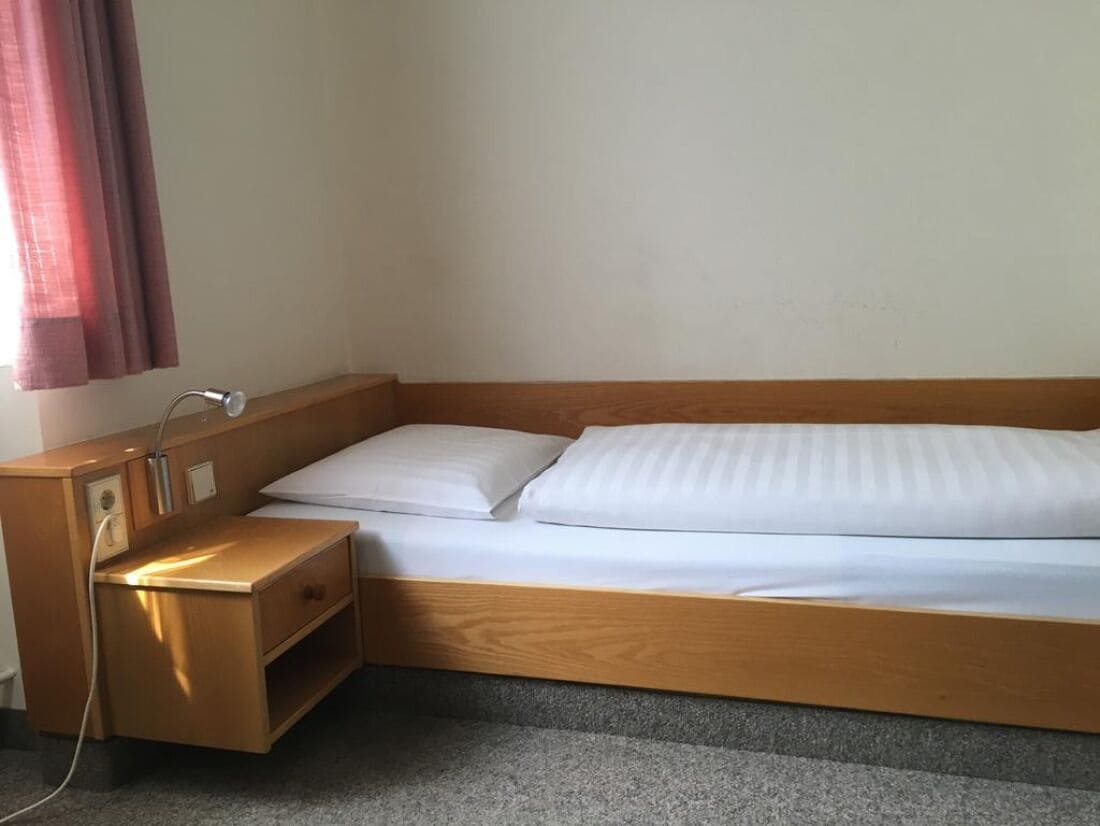 A single bed with a wooden frame and white bedding, a bedside table with a lamp, and a power outlet.