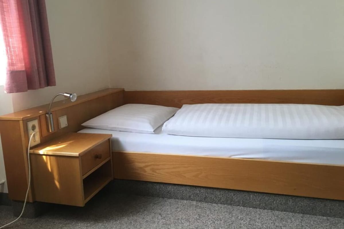 Una cama individual con estructura de madera y ropa de cama blanca, una mesita de noche con lámpara y un enchufe.