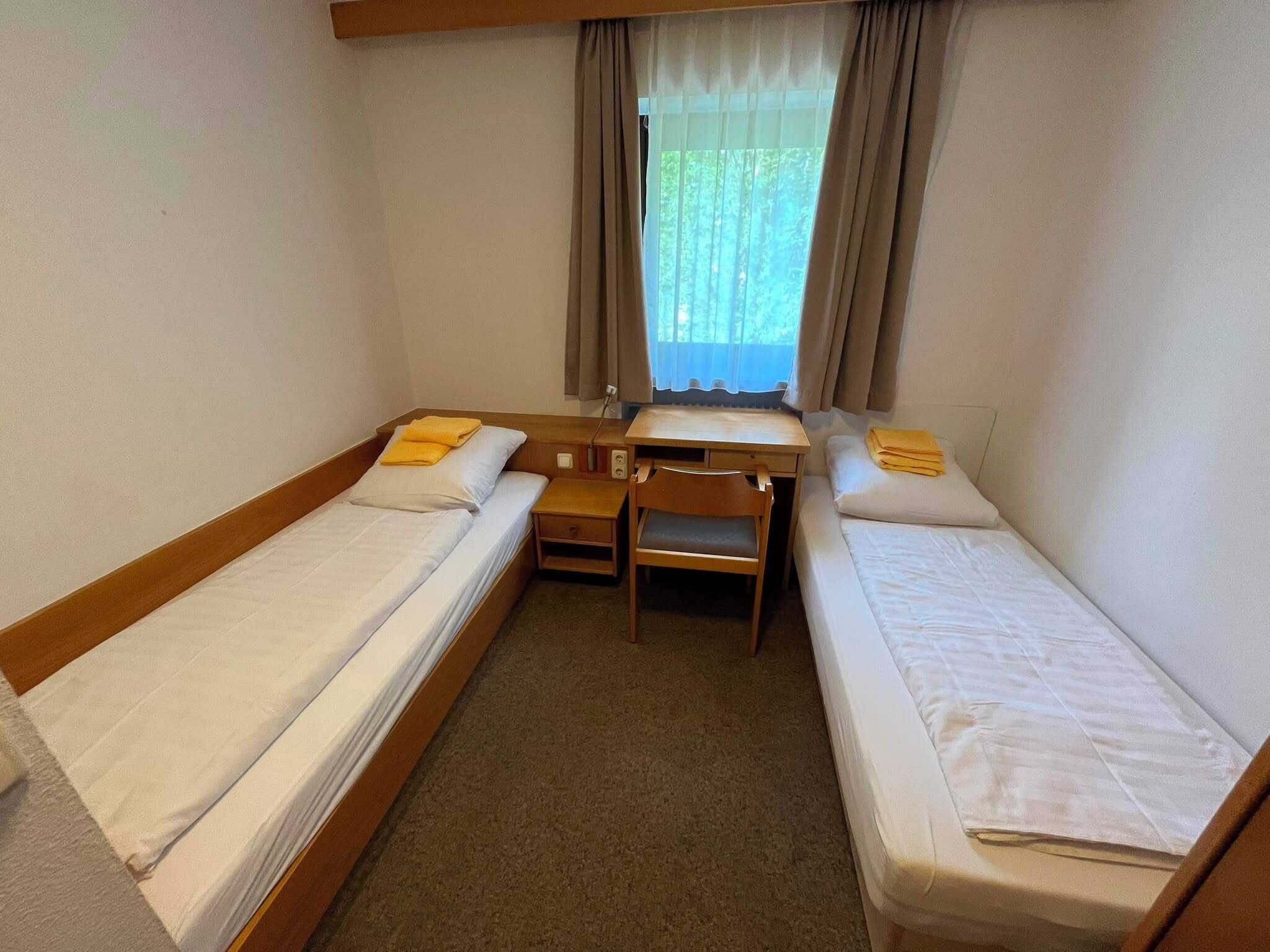 A small, compact room with two single beds, a desk, and a chair.