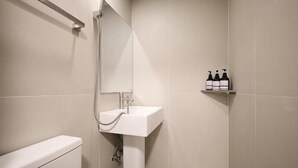 Un baño con un lavamanos blanco, un espejo y una ducha. Hay tres botellas en un estante.