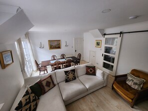 A living room with a sofa, a dining table, and chairs. There are two doors, one leading to a balcony and another to a bedroom.