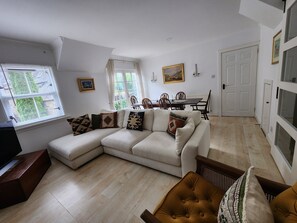A living room with a sectional sofa, a dining table with chairs, a television, and a window with blinds.