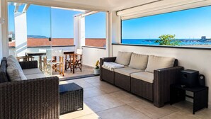 A balcony with a wicker sofa, a small table, and chairs, overlooking a coastal view with boats and buildings. - Huge Terrace Sea Front (Bosa Marina)