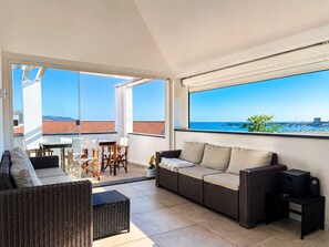 Ein Balkon mit Rattan-Sofa, einem kleinen Tisch und Stühlen mit Blick auf die Küste, wo Boote und Gebäude liegen.