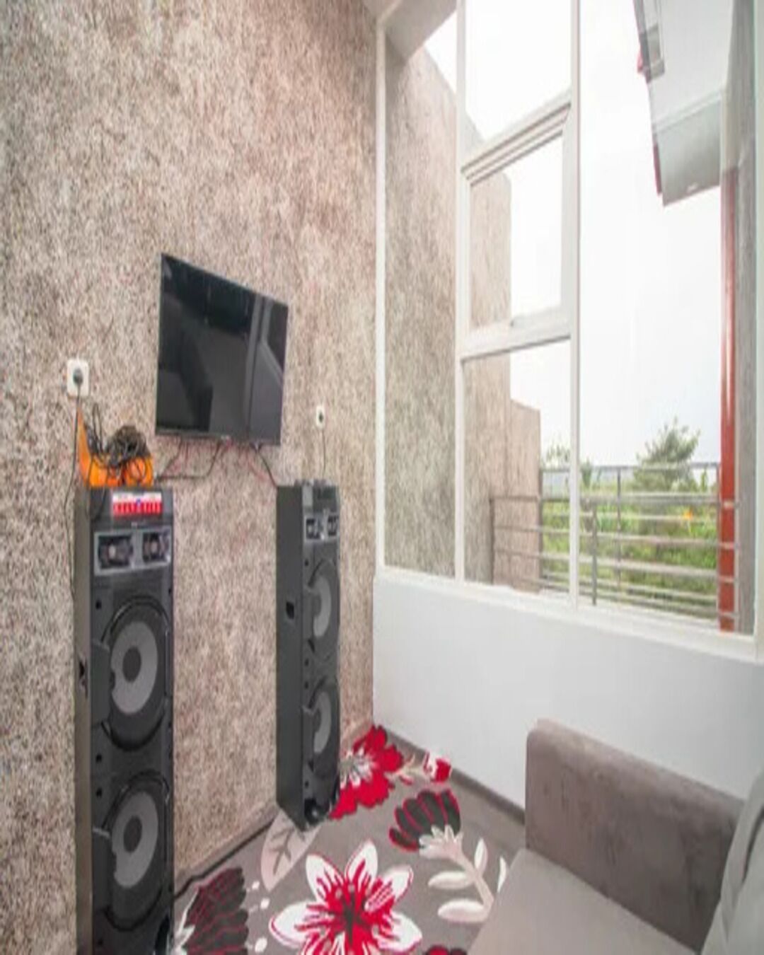 A room with a television mounted on the wall, two speakers, a window with a view, and a floral carpet.