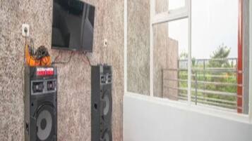 A room with a television mounted on the wall, two speakers, a window with a view, and a floral carpet.