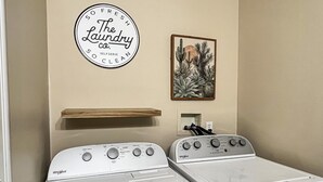 A laundry room with two Whirlpool washers, a wooden shelf, a framed cactus artwork, and a circular sign on the wall. - Quiet 1st-Floor 2BR Haven w/ King Bed, Desks, In-Unit Laundry & 2-Car Parking | Walk to Yale (New Haven)