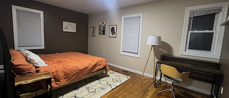 A bedroom with a bed, a desk, and a chair. There are two windows with blinds, a lamp, and a rug on the floor.