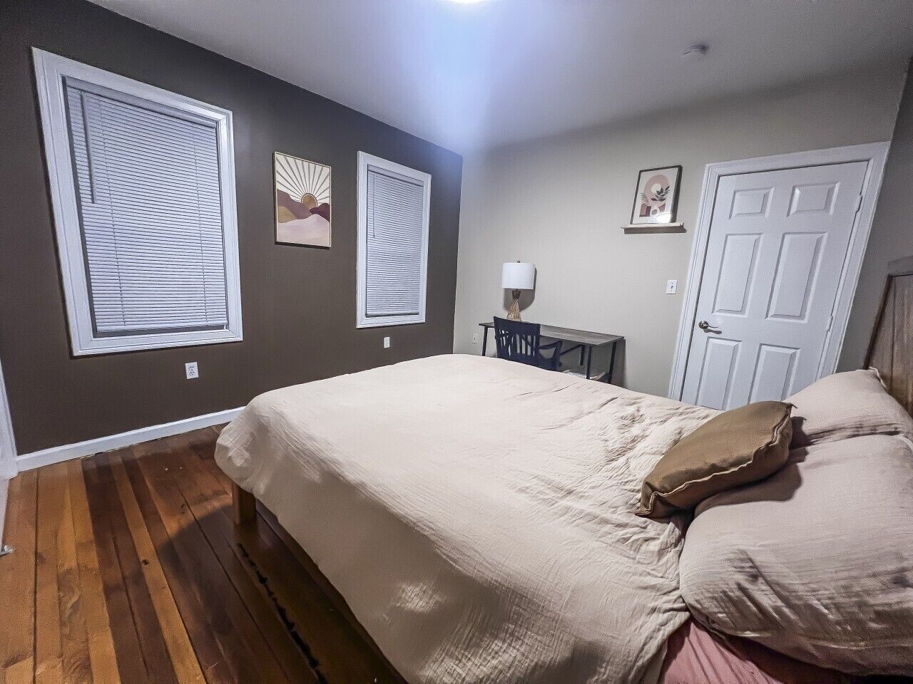 A bedroom with a bed, a desk, and a chair. There are two windows with blinds, a door, and a wall-mounted lamp.