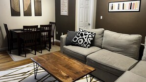 A living room with a sofa, coffee table, dining table, chairs, and artwork on the walls. - Quiet 1st-Floor 2BR Haven w/ King Bed, Desks, In-Unit Laundry & 2-Car Parking | Walk to Yale (New Haven)
