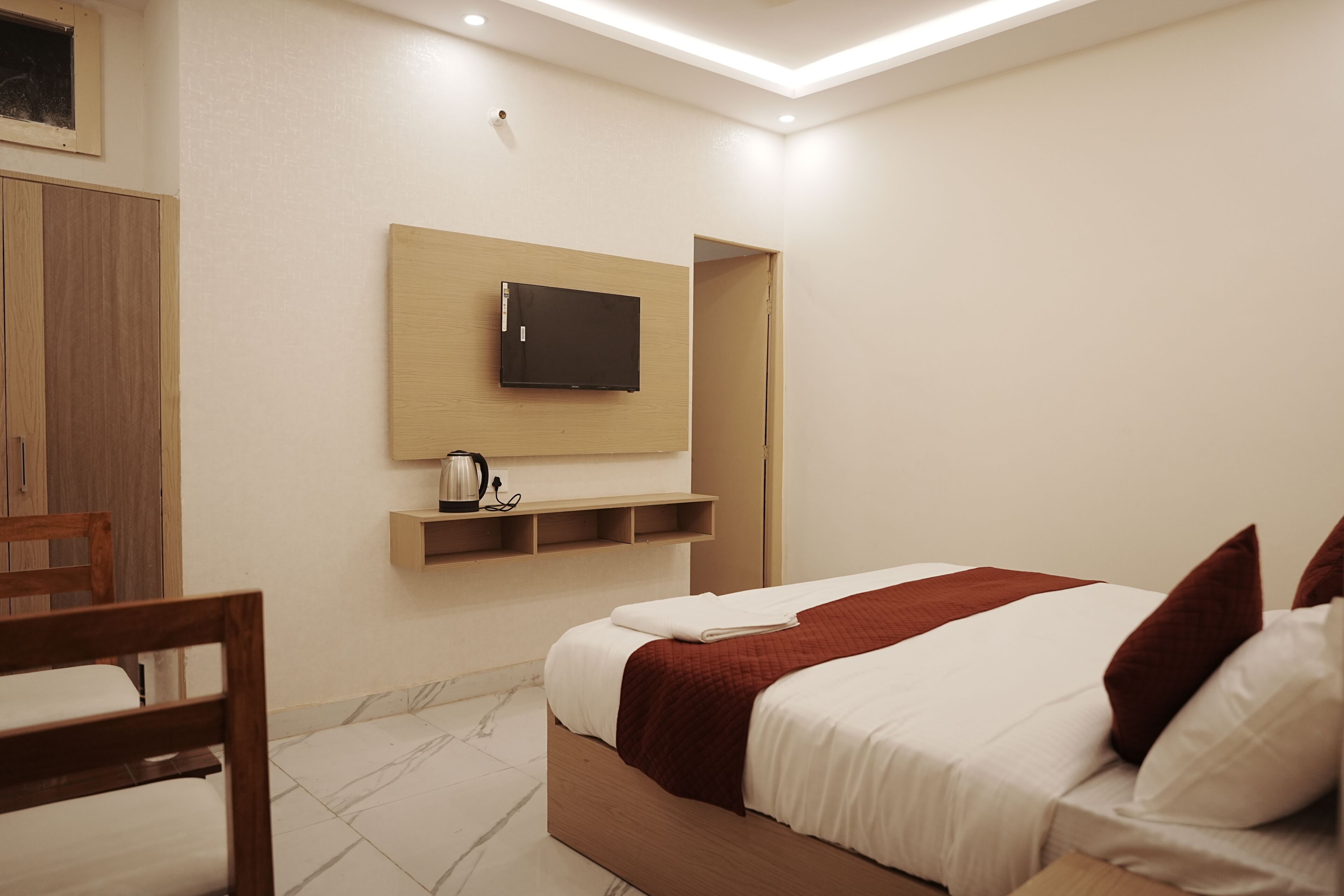 A hotel room with a bed, a TV mounted on the wall, a wooden chair, and a shelf with a kettle.