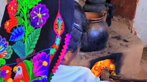 Una persona in abiti tradizionali con un elaborato copricapo ricamato, in piedi davanti a una pentola sul fornello.
