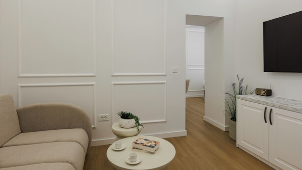 Een moderne woonkamer met een bank, een rond bijzettafeltje met kopjes en een boek, een kleine plant op een voetstuk, en een aan de muur bevestigde televisie.