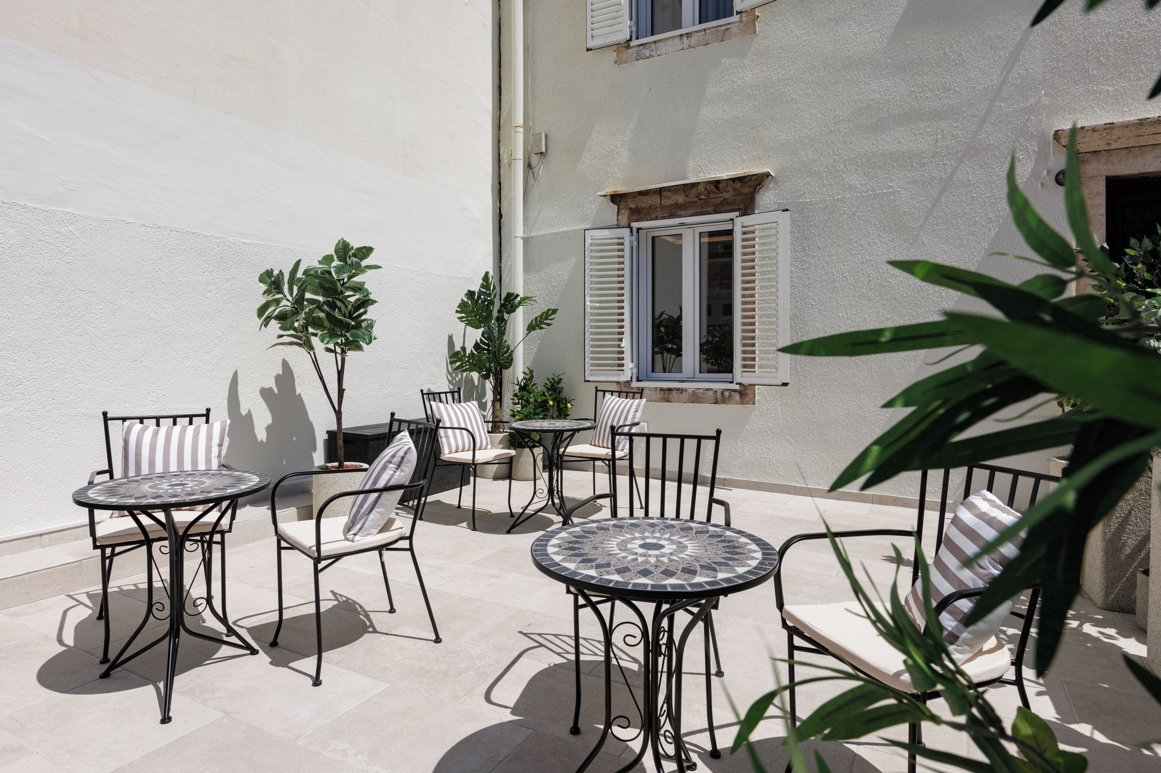Un patio al aire libre con varias mesas y sillas redondas, rodeado de plantas en macetas y un edificio de paredes blancas y una ventana con contraventanas.