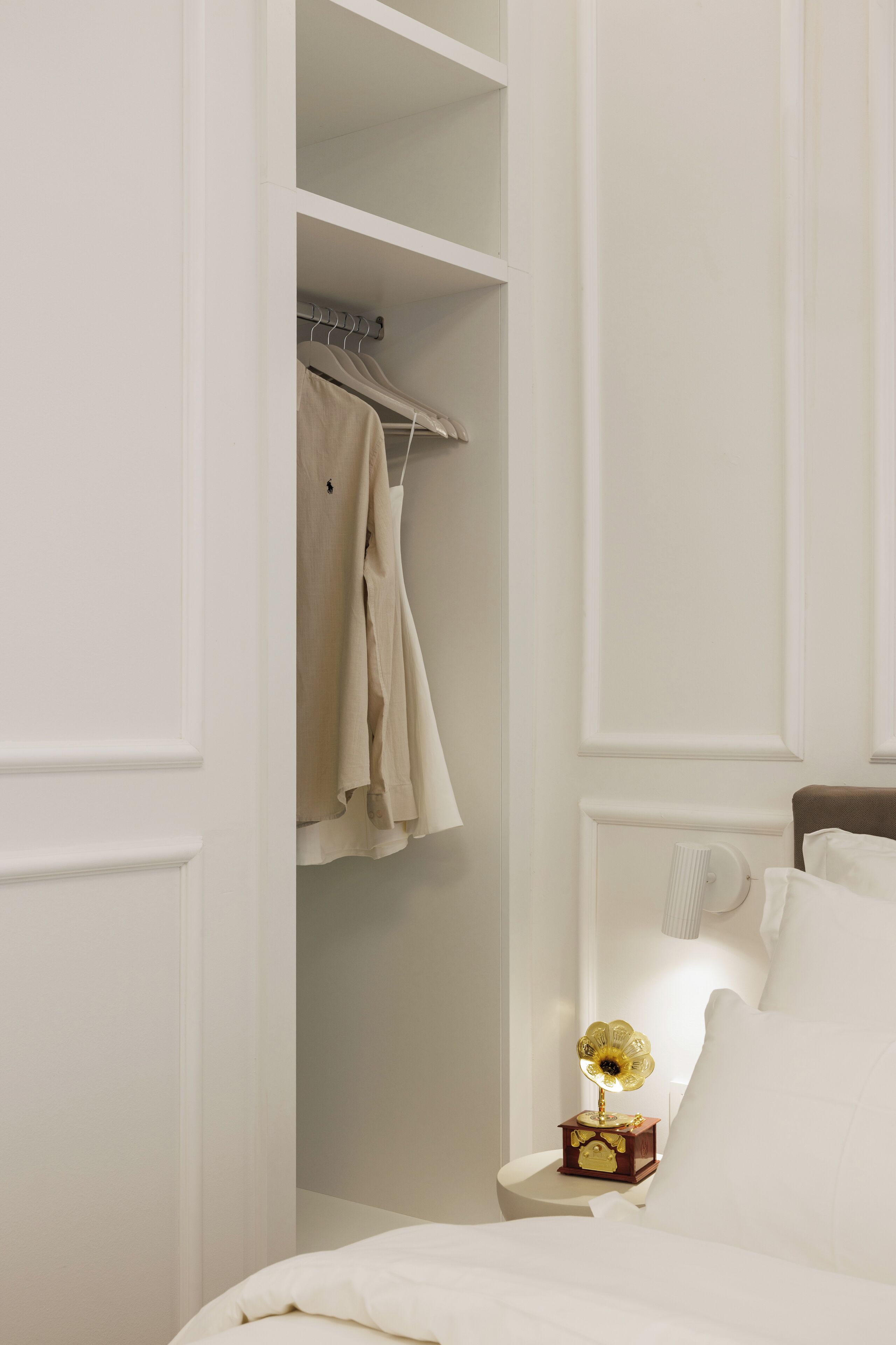 Un dormitorio con una cama, una mesita de noche con un gramófono y un armario con ropa colgada.