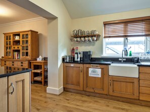 A kitchen with wooden cabinets, a coffee station, and a window with blinds. - St Davids Meadow House (Woodford)
