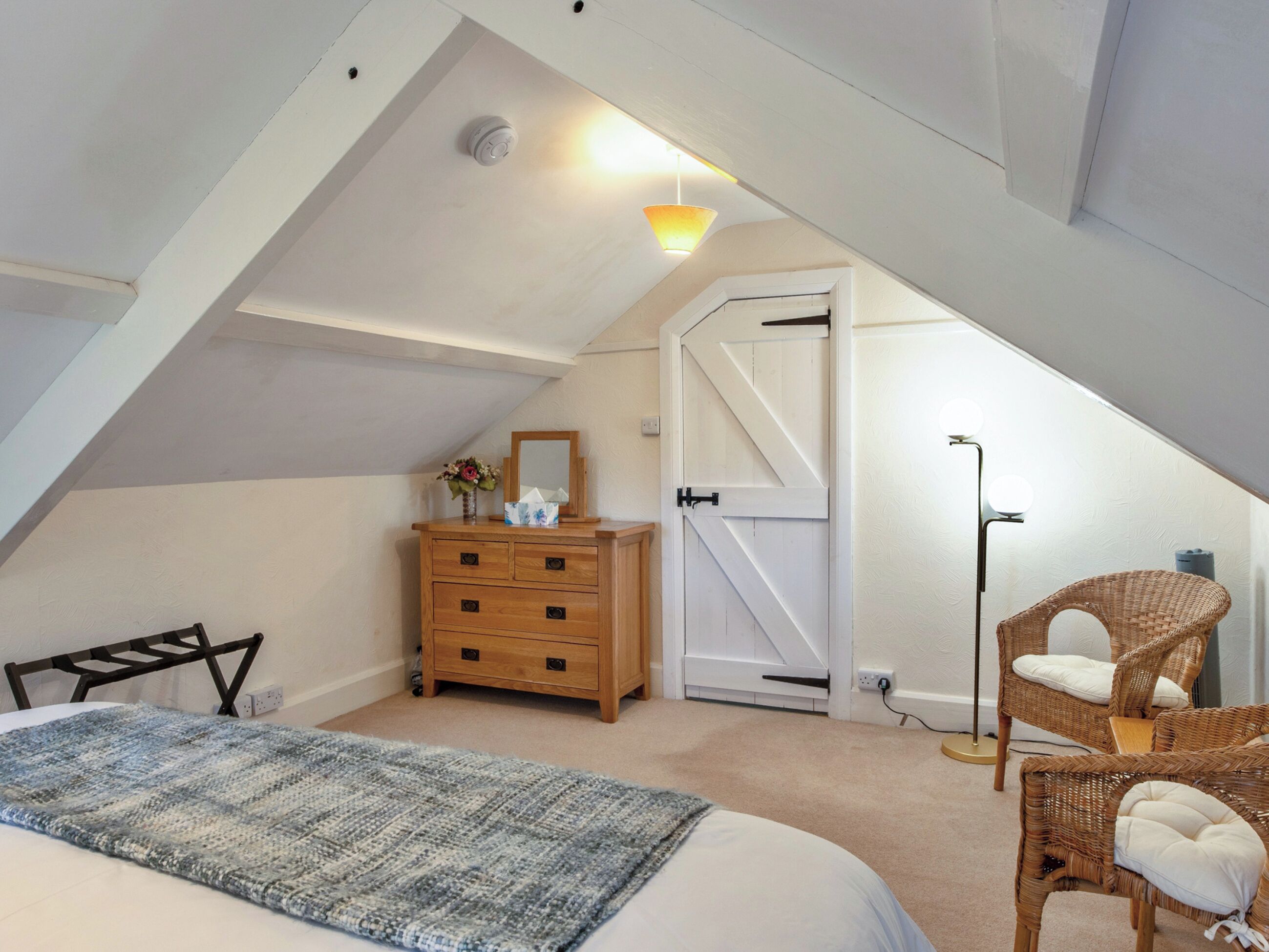Une chambre avec un plafond en pente, un lit, une commode, un miroir, une porte et un lampadaire.