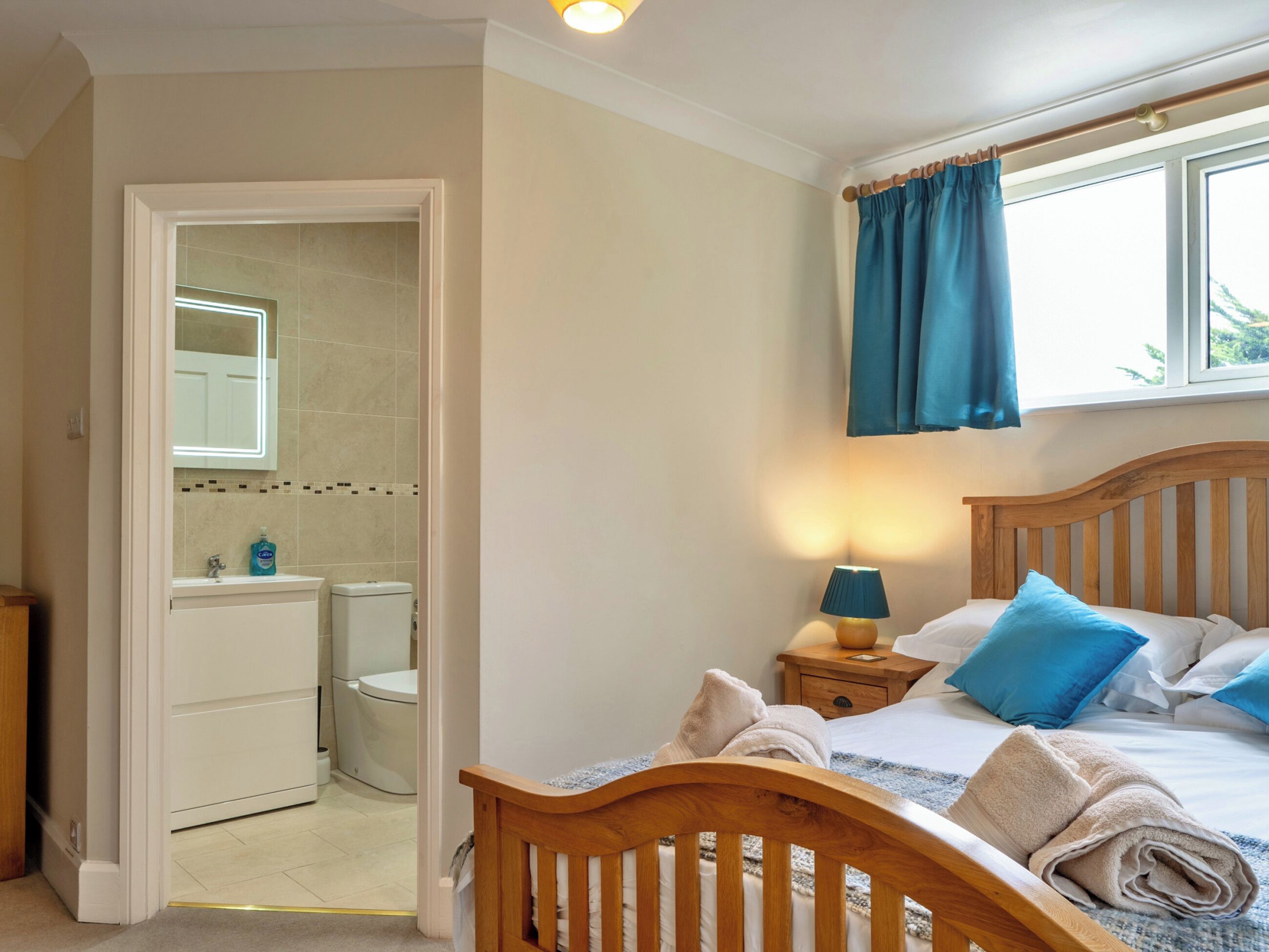 Ein Schlafzimmer mit einem hölzernen Bett, einem Nachttisch mit Lampe, einem Fenster mit Vorhängen sowie einem Badezimmer mit Toilette und Waschbecken.
