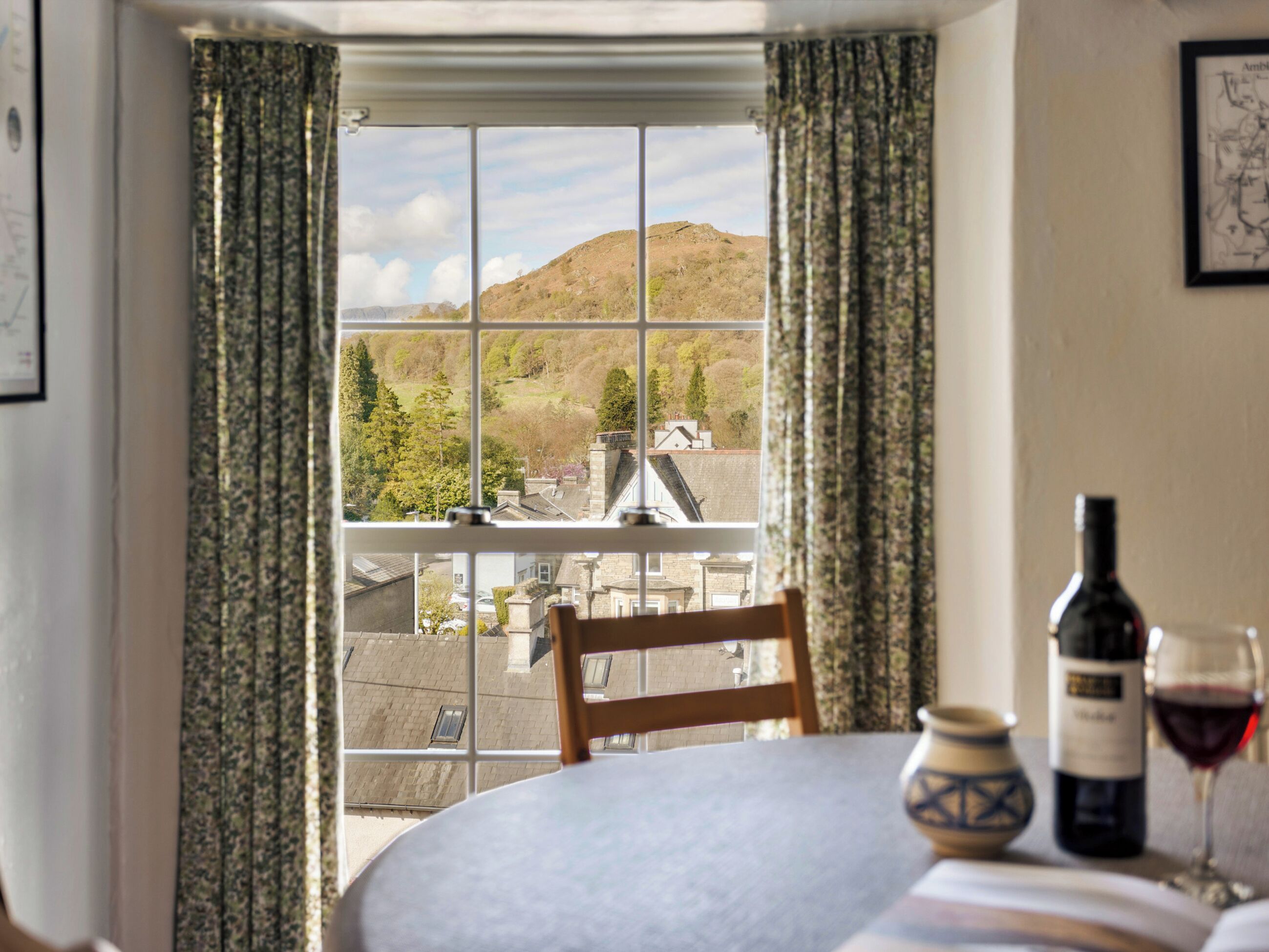 A dining area with a table set for two, a window with a view of a hillside, and a bottle of wine on the table.
