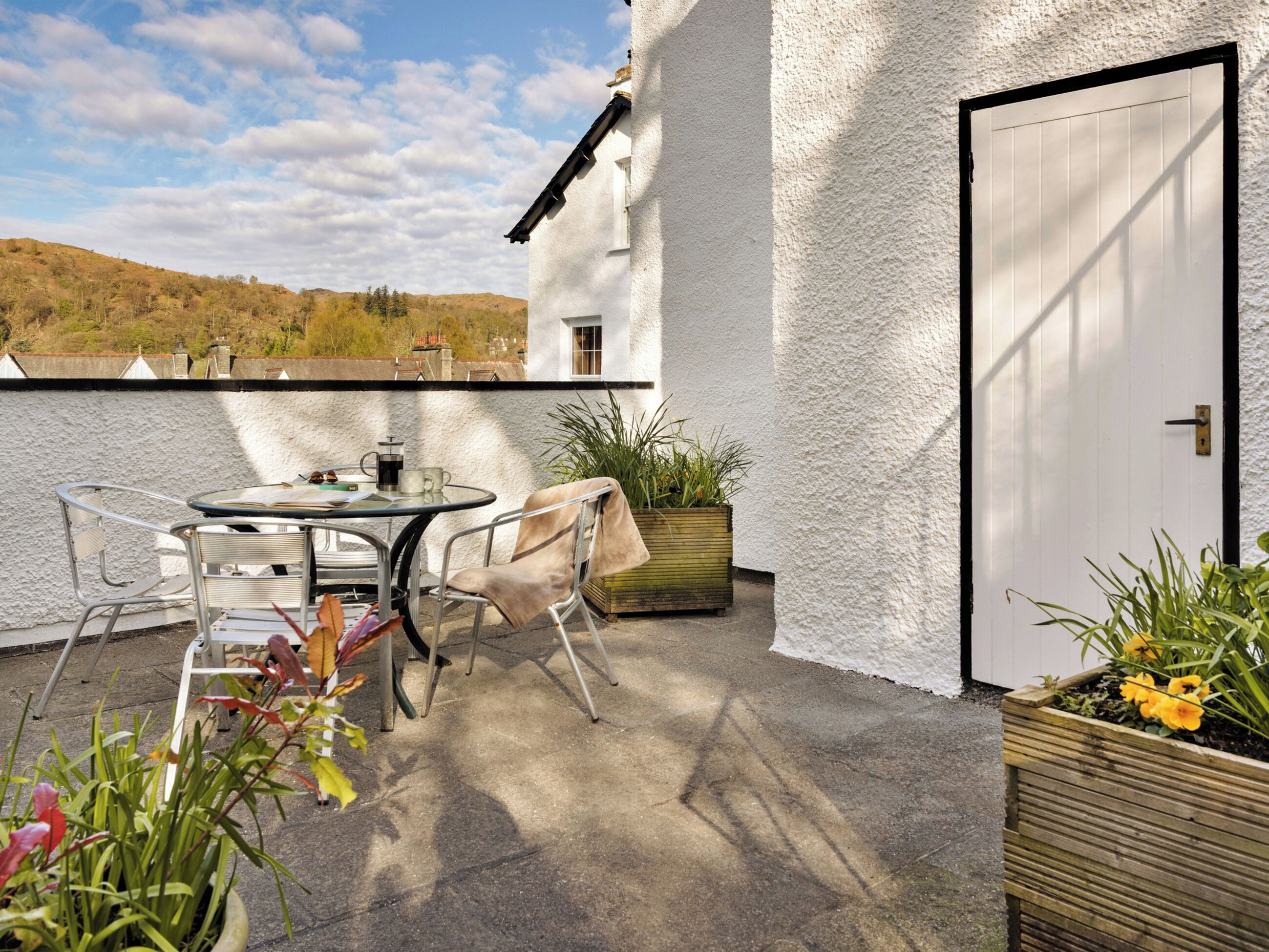 Une terrasse avec une table et des chaises, un jardinier en bois avec des plantes et une porte blanche.