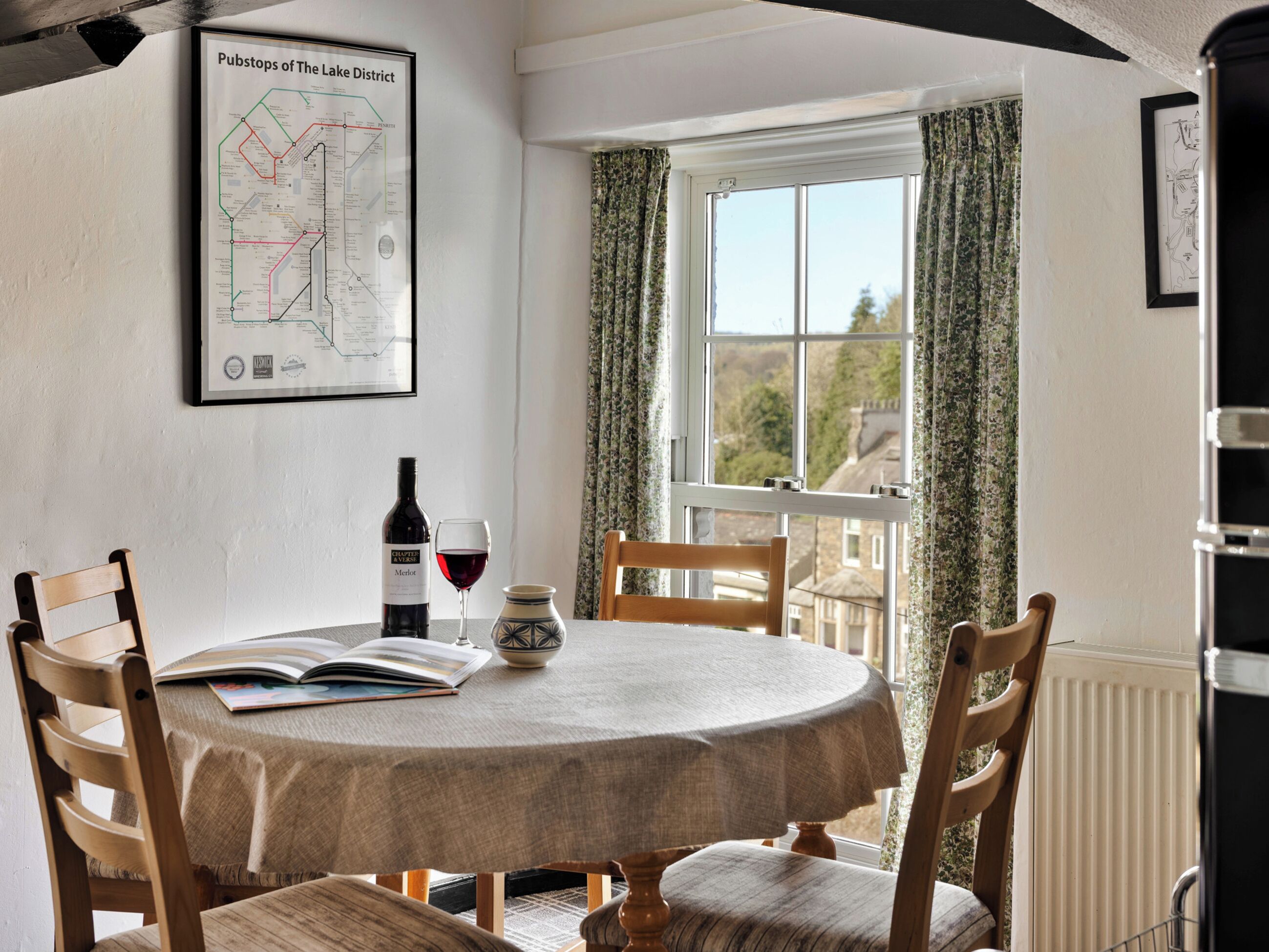A dining area with a round table, chairs, a wine bottle, and a glass of red wine. A map titled 'Pubstops of The Lake District' is on the wall.