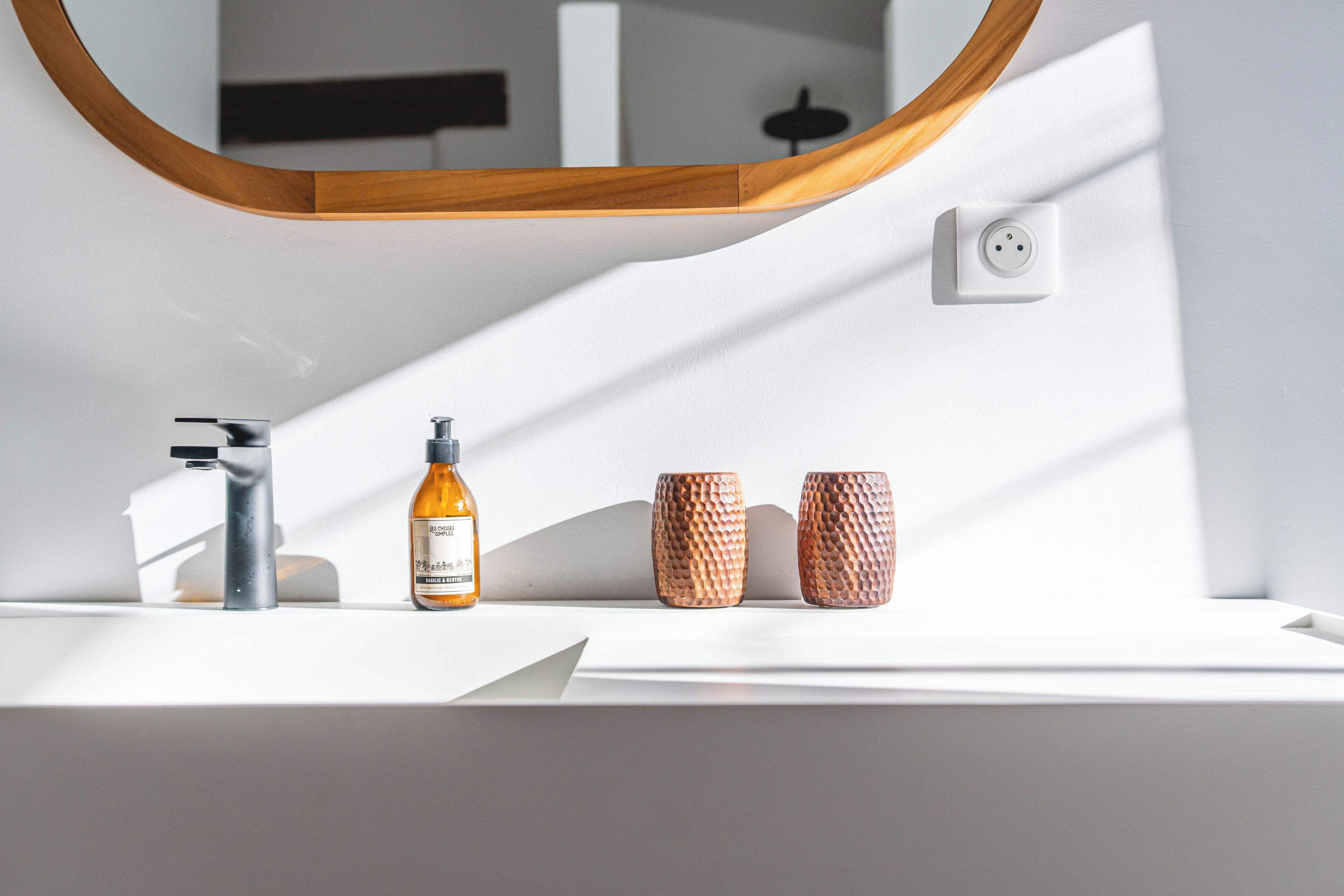 Une salle de bain avec un lavabo blanc, un miroir, un flacon de savon liquide et deux tasses à motifs.