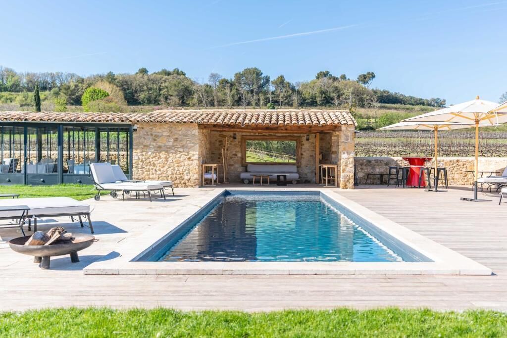 Piscina con sillas de descanso y una pérgola, rodeada de muros de piedra y un viñedo al fondo.