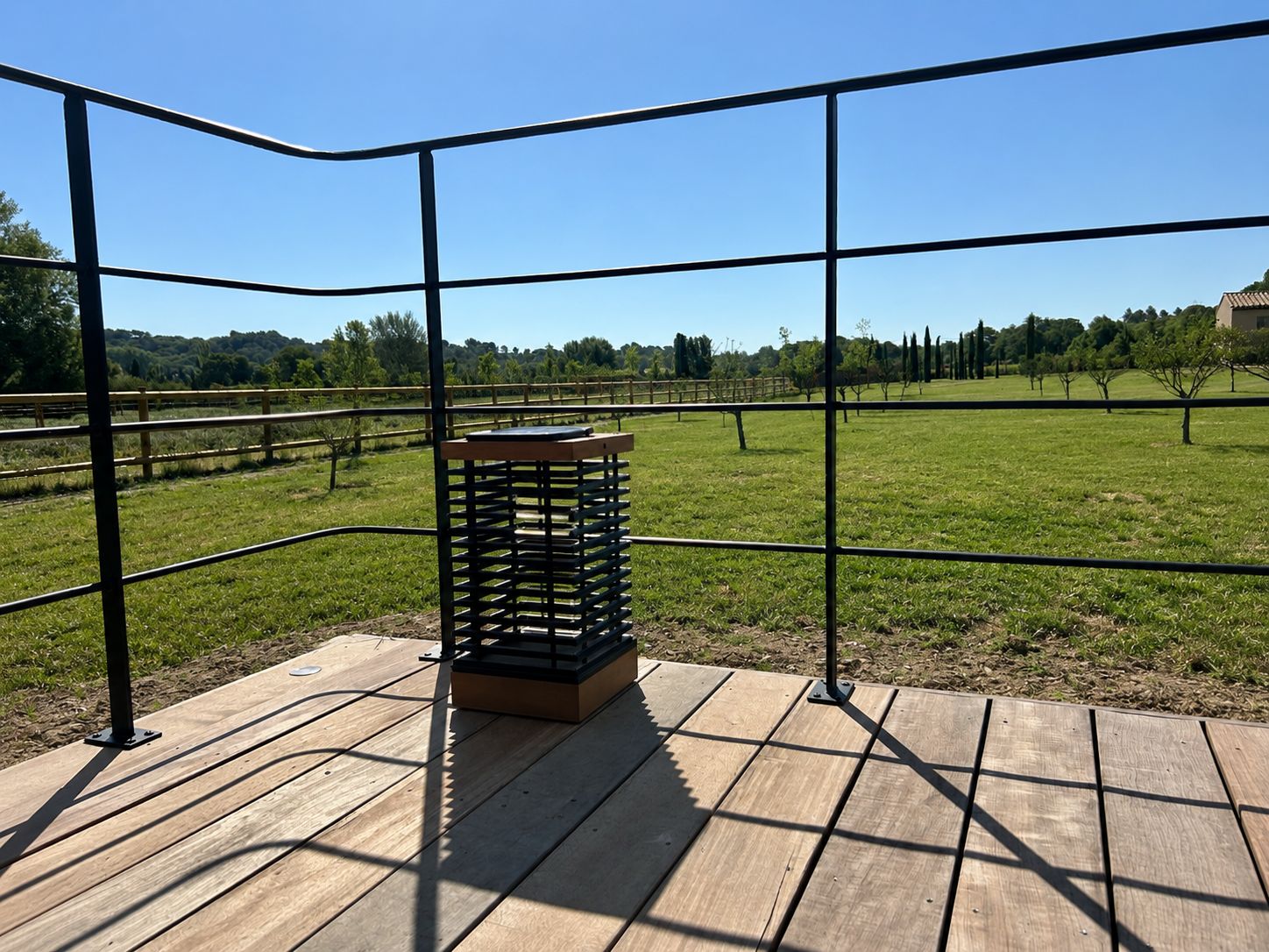 Eine Holzterrasse mit Metallgeländer, die auf ein Wiesfeld mit Bäumen und ein Gebäude in der Ferne blickt.