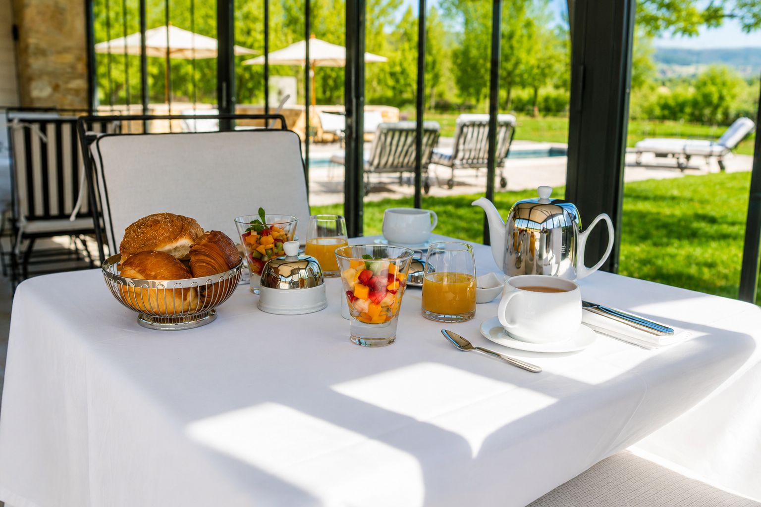 Mesa de café da manhã com croissants, frutas, café e chá, com vista para uma piscina e jardim.