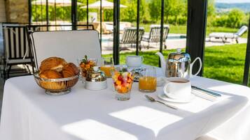 Mesa de café da manhã com croissants, frutas, café e chá, com vista para uma piscina e jardim.