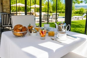 A breakfast table with croissants, fruit, coffee, and tea set, overlooking a pool and garden.